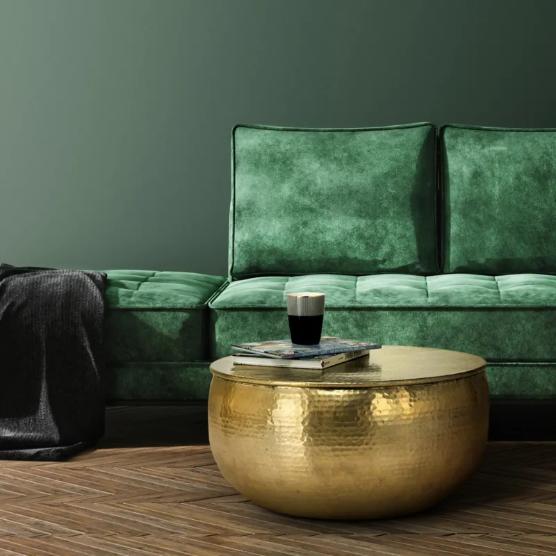 Grünes Sofa mit Kissen und einem runden goldenen Tisch, darauf ein Glas und Bücher.